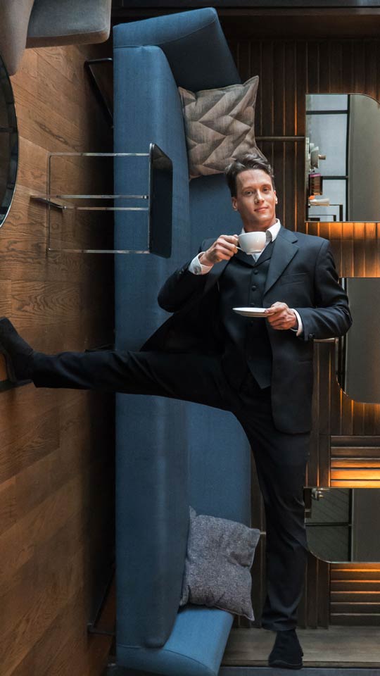 Man in black three piece suit and waistcoast with black dress trousers holding a tea cup drinking vertically horizontally in an elegant lounge